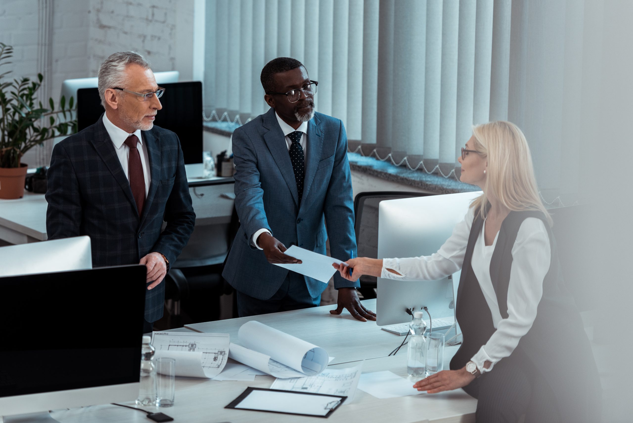 african american businessman giving blank paper to attractive blonde woman near partner in office