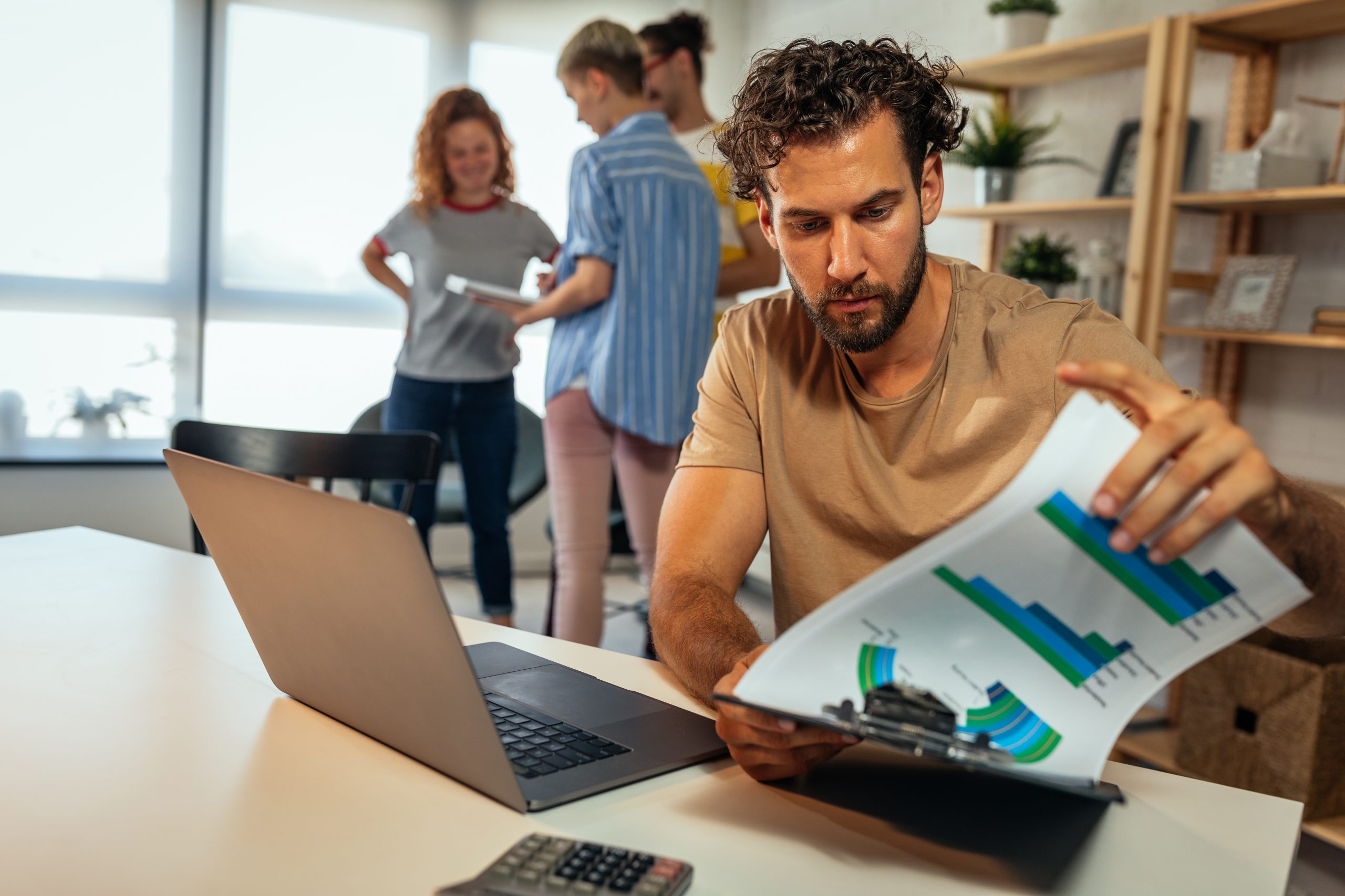 Young man working on new project while his coworkers standing behind him and conversing